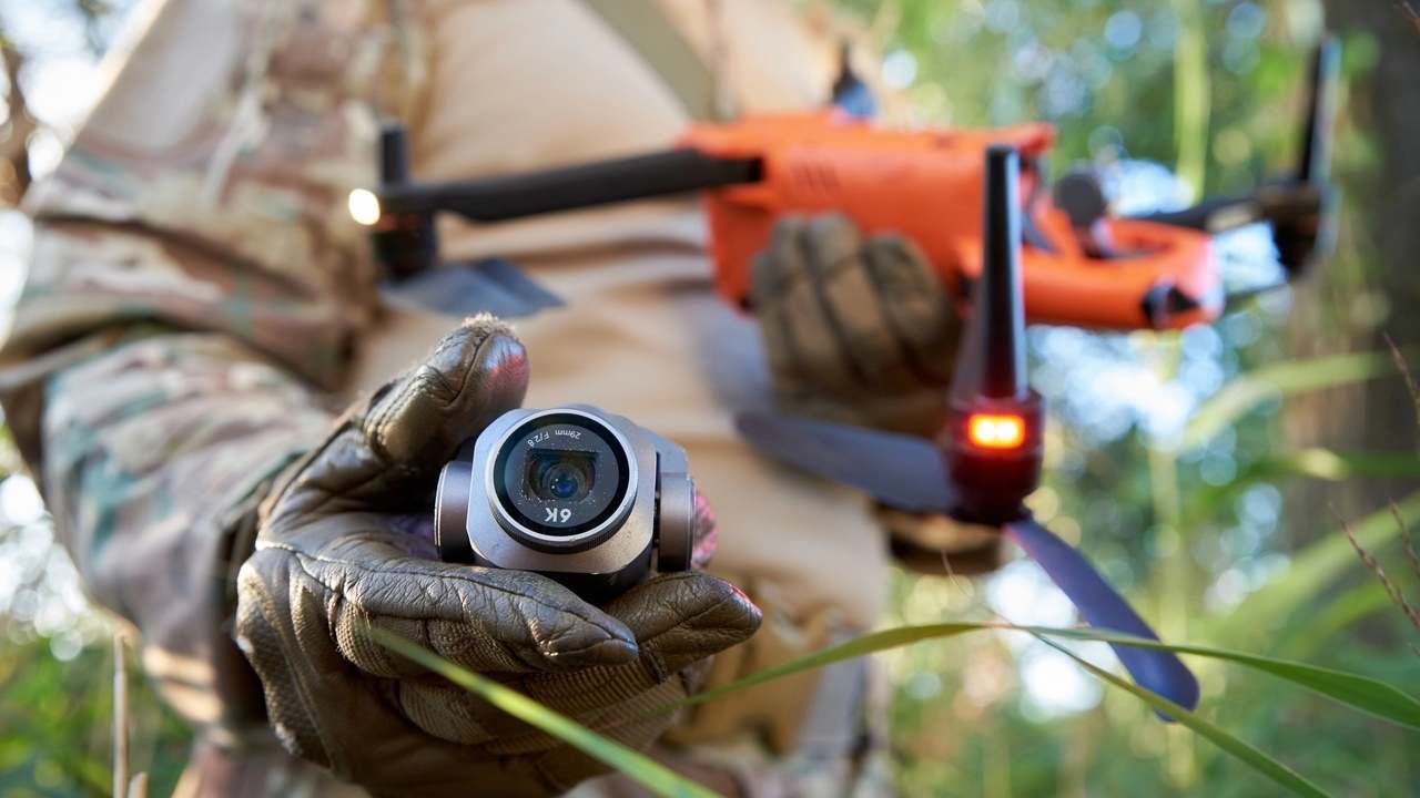 Vojak v maskovacej uniforme drží malý prieskumný dron, zatiaľ čo v pozadí je pripravený väčší bezpilotný systém.