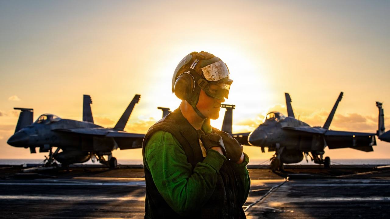 Člen posádky na palube lietadlovej lode USS Gerald R. Ford pri západe slnka s lietadlami v pozadí